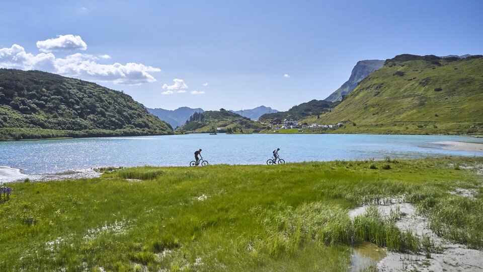 Zwei Radfahrer fahren entlang eines glitzernden Sees. Im Hintergrund sind grüne Hügel und ein blauer Himmel zu sehen.