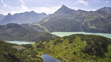 Eine atemberaubende Berglandschaft mit grünen Hügeln und einem klaren See. Im Hintergrund erheben sich majestätische Berge unter einem blauen Himmel.