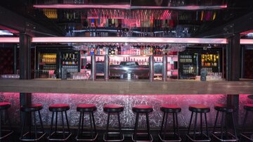 A stylish bar with a long wooden counter and modern seating. The lighting is in soft pink tones, creating an inviting atmosphere.