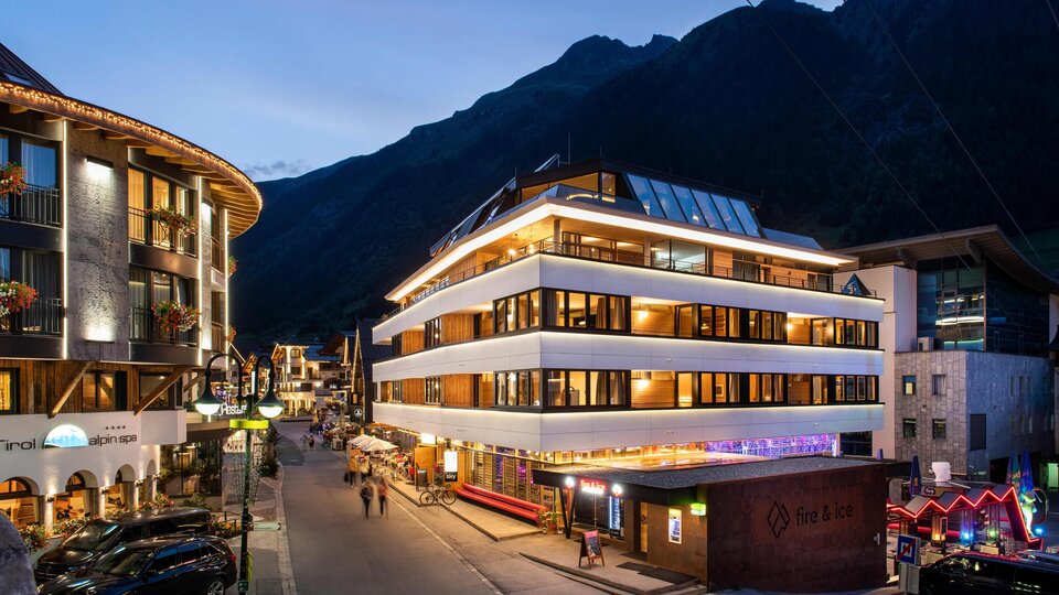 A modern building in a picturesque alpine town at twilight. In the background, there are high mountains and a busy street with lights.