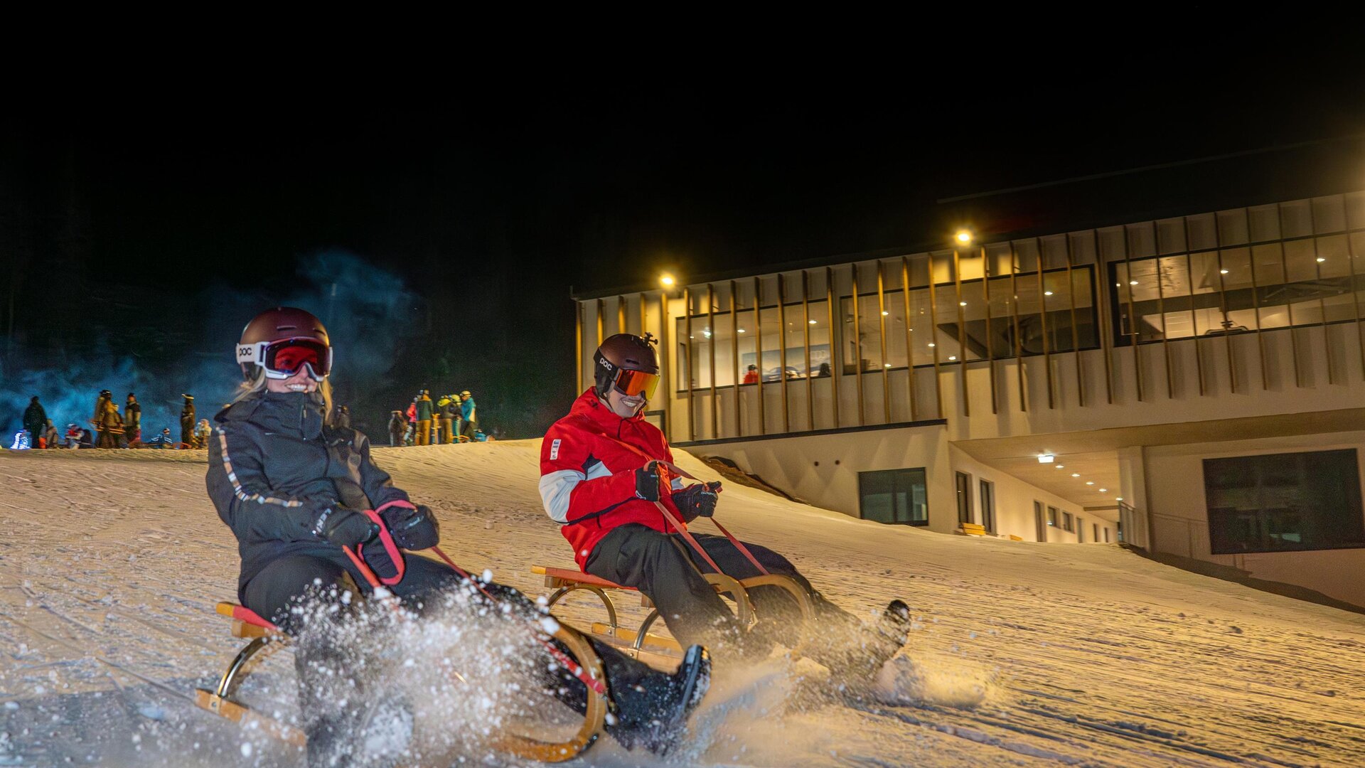 Two people are riding a sled on a lit snow slope at night. In the background, a building can be seen.