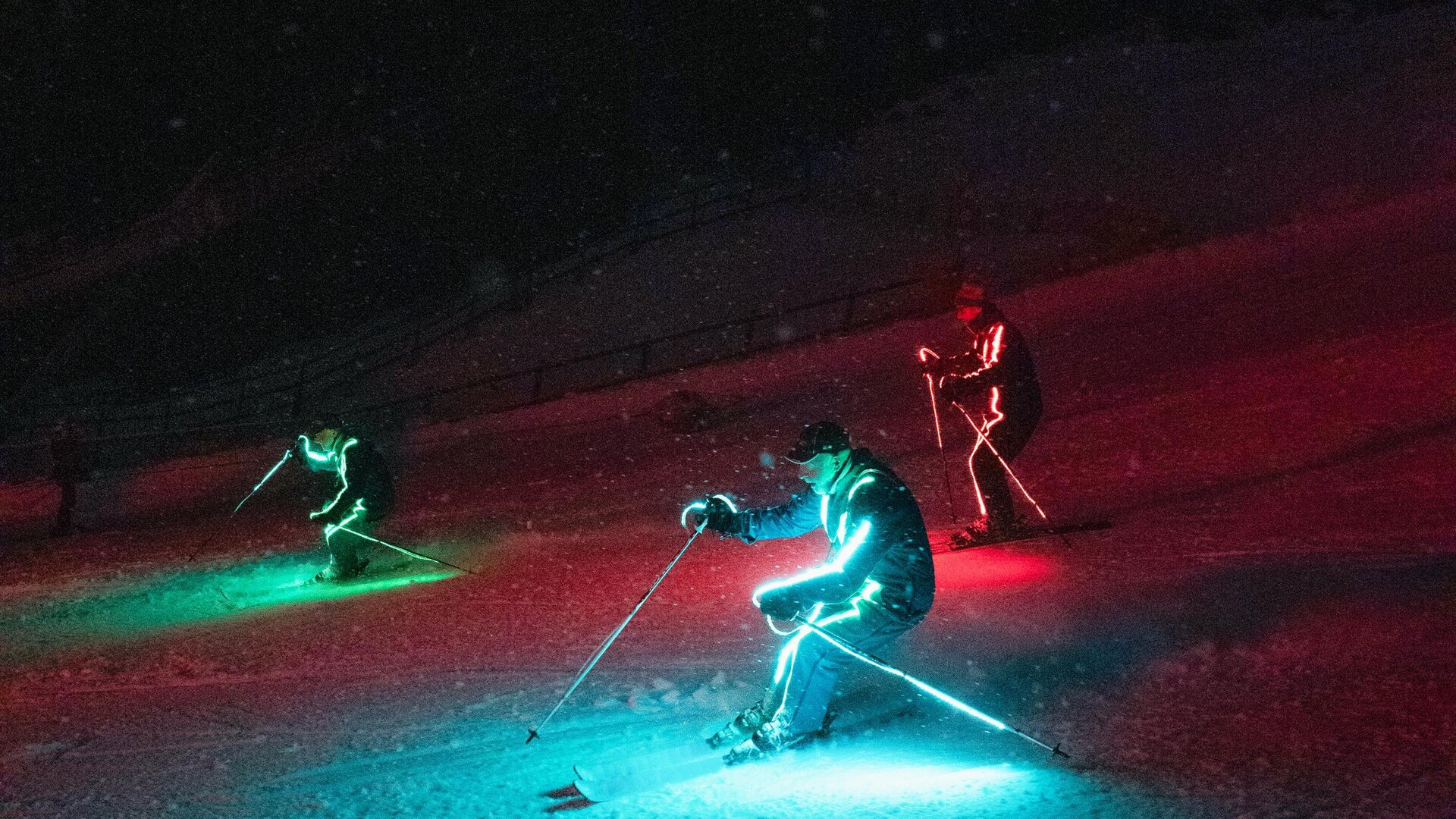 Three skiers with bright, colorful lights descend a slope in the dark. The surroundings are covered in snow and the atmosphere is festive.
