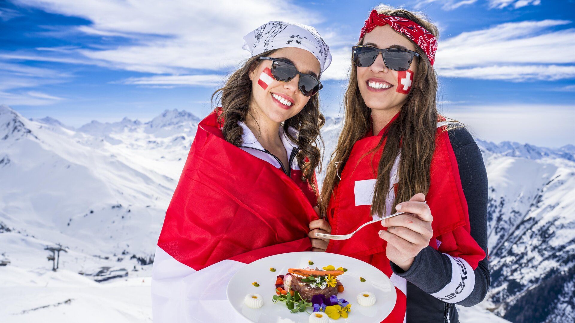 Zwei Frauen in Sonnenbrillen stehen auf einem schneebedeckten Berg und halten ein Tablett mit Essen. Sie tragen Fahnen von der Schweiz und lächeln vor einer beeindruckenden Berglandschaft.