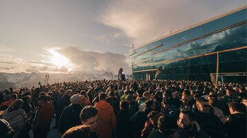 A large crowd gathers in front of a futuristic building in the mountains. In the background, snow-capped peaks and a sunset can be seen.