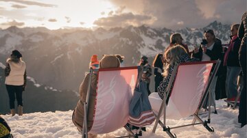 Two people are sitting relaxed in lounging chairs on the snow and enjoying the view of the mountains. In the background, other people can be seen also appreciating the scenery.