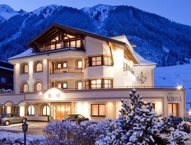 Ein charmantes Hotel im Schnee mit einem malerischen Bergpanorama im Hintergrund. Die Fassade ist hell erleuchtet und strahlt eine einladende Atmosphäre aus.