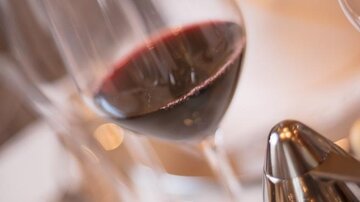 Ein elegantes Weinglas mit rotem Wein steht auf einem Tisch. Im Hintergrund sind weitere Gläser und Besteck zu sehen.