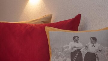 Ein gemütliches Bett mit roten Kissen und einem dekorativen Kissen mit einem Schwarz-Weiß-Foto von zwei Frauen beim Skifahren. Eine Wandlampe sorgt für sanftes Licht.