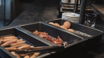 Ein Buffet mit verschiedenen Frühstückszutaten. Es gibt Würstchen, Speck, Eier und Gewürze.