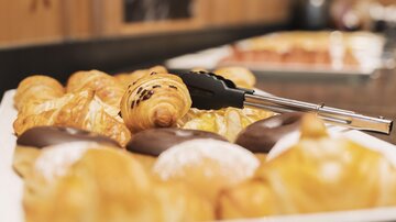 Eine Auswahl von köstlichen Backwaren, darunter Croissants und Schokoladendoughnuts. Im Hintergrund sind weitere Leckereien zu sehen.