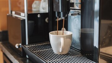 Eine moderne Kaffeemaschine, die frischen Kaffee in eine weiße Tasse einschenkt. Im Hintergrund sind einige Tassen und die Maschine selbst sichtbar.