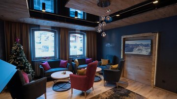 Eine gemütliche Lounge mit modernen Möbeln und warmen Farbtönen. Im Raum steht ein Weihnachtsbaum, und große Fenster lassen viel Tageslicht hinein.