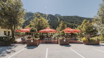 Ein gemütlicher Außenbereich mit roten Sonnenschirmen und Stühlen. Im Hintergrund sind die grünen Berge zu sehen.