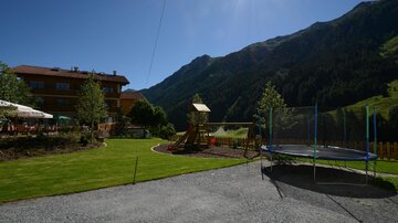 Ein schöner Garten mit einem Spielplatz und einer Trampolin. Im Hintergrund sind grüne Berge und ein blauer Himmel zu sehen.