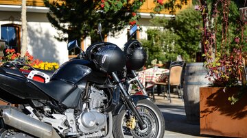 Eine schwarze Motorrad steht in einem gemütlichen Außenbereich mit Blumen und Tischen. Im Hintergrund sind Gäste zu sehen, die sich entspannen.