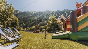 Ein schöner Spielplatz mit einer großen Hüpfburg und einem atemberaubenden Blick auf die Berge. Im Vordergrund stehen Liegestühle auf der grünen Wiese.