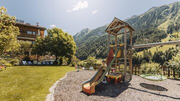 Ein Spielplatz mit einer Rutsche steht im Vordergrund. Im Hintergrund sind schöne Berge und eine grüne Wiese zu sehen.