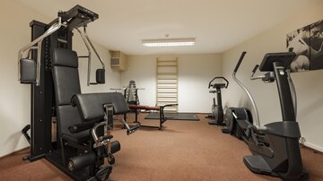 Ein modernes Fitnessstudio mit verschiedenen Trainingsgeräten. Der Raum ist gut beleuchtet und hat einen Teppichboden.