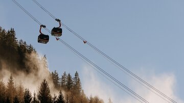 Eine Seilbahn fährt über bewaldete Hügel im Nebel. Der klare Himmel und die sanften Berge schaffen eine friedliche Atmosphäre.