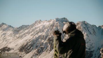 Ein Skifahrer hebt seine Skier vor einer beeindruckenden Berglandschaft. Die schneebedeckten Berge strahlen im klaren blauen Himmel.