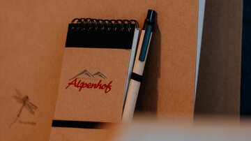 Ein Notizblock mit dem Logo "Alpenhof" und ein schwarzer Stift liegen auf einem Tisch. Im Hintergrund sind braune Ordner sichtbar.