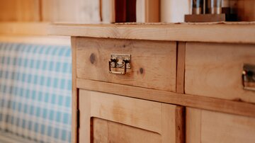 Eine rustikale Holzkommode mit Schubladen und einem dekorativen Element. Im Hintergrund ist eine gepolsterte Sitzbank mit kariertem Muster zu sehen.
