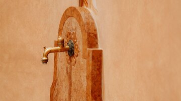 Ein dekorativer Wasserhahn aus Messing, der an einer kunstvollen Wand installiert ist. Die Wand hat eine warme, orangefarbene Oberfläche.