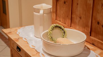 Eine weiße Schüssel mit einer dekorativen Frucht und einem Kranz aus Blumen steht auf einem Holztablett. Daneben befindet sich ein weißer Krug auf einer hellen Unterlage.