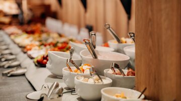 Ein reichhaltiges Buffet mit verschiedenen Speisen in weißen Schalen. Im Hintergrund sind frische Früchte und andere Gerichte sichtbar.