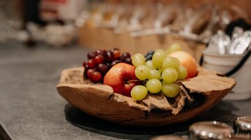 Eine Schale mit frischem Obst, darunter Äpfel, Trauben und grüne Weintrauben. Der Hintergrund ist unscharf und zeigt eine gemütliche Umgebung.