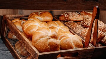 Ein Korb mit frischen Brötchen, die goldbraun gebacken sind. Im Hintergrund sind verschiedene Brötchen mit Sesam und anderen Toppings zu sehen.