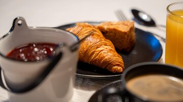 Ein Frühstückstisch mit einem Croissant, einem Stück Kuchen und Marmelade. Dazu gibt es Kaffee und Orangensaft.