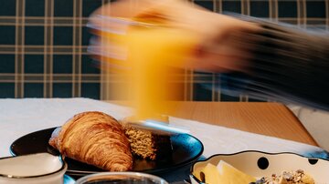 Ein Frühstückstisch mit einem Croissant, einem Stück Kuchen und einer Schüssel Müsli. Im Hintergrund wird ein Glas Orangensaft gehalten.