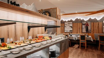 Ein Buffet mit einer Auswahl an Speisen, darunter frisches Gemüse und Obst, in einem modernen Restaurant. Der Raum ist hell und einladend, mit Holz- und Steinelementen dekoriert.
