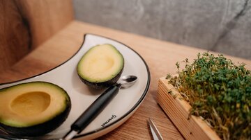 Eine geschnittene Avocado auf einem teller mit einem silbernen Löffel. Daneben befindet sich ein kleines Holzgefäß mit frischen Sprossen.