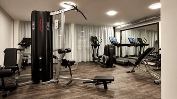 A modern gym with various equipment. The room is bright and well-lit, with large windows and wooden flooring.