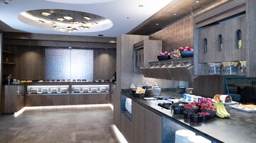 A modern kitchen with elegant wooden paneling and a variety of breakfast offerings. Fresh fruits, pastries, and drinks are arranged on the counter.
