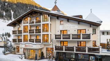 Ein charmantes Hotel im Alpenstil, umgeben von schneebedeckten Bergen. Große Fenster lassen warmes Licht ins Innere strömen.
