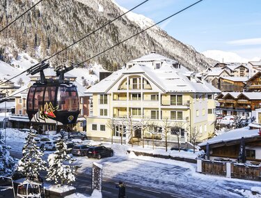 Eine malerische Winterlandschaft mit schneebedeckten Bergen und einem Seilbahnzug. Im Vordergrund ist ein charmantes Gebäude sichtbar.