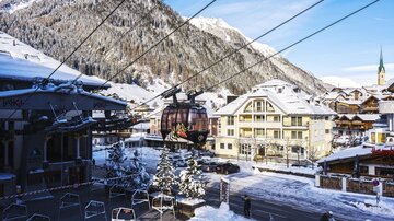 Eine schneebedeckte Berglandschaft mit einem Skilift und traditionellen Chalets. Der Himmel ist klar und blau, und es gibt Bäume mit Schnee.