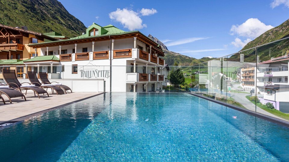 A modern hotel with a beautiful pool and loungers. In the background, green mountains and a clear sky can be seen.