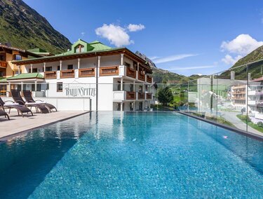 A modern hotel with a beautiful pool and loungers. In the background, green mountains and a clear sky can be seen.