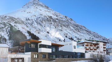 A modern building in the foreground, surrounded by snow-covered mountains. The clear blue sky completes the winter landscape.