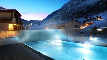 A beautiful outdoor pool in the mountains at dusk. The lights reflect on the water and the snow-covered mountains are visible in the background.