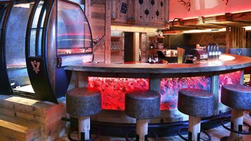 A modern bar with wooden design and cozy seating. The atmosphere is highlighted by colored lighting.