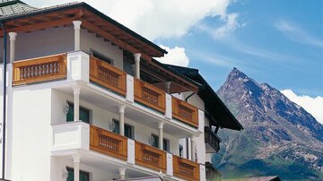 A modern building with wooden balconies and a beautiful mountain landscape in the background. The sky is clear and the surroundings are picturesque.