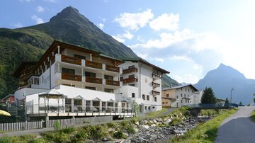 A modern building in the mountains, surrounded by green nature. The sky is clear and the surroundings radiate tranquility.