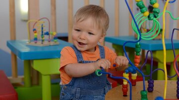 A small child with blonde hair is playing at a colorful play table. They are smiling and moving the beads on a shape toy.
