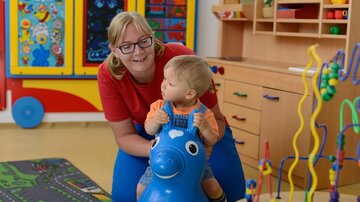 A happy child is riding on a blue toy horse while a caregiver smiles. In the background, colorful play equipment and a rug with road motifs can be seen.