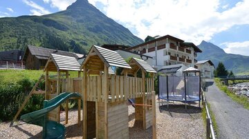 A wooden playground with a slide and trampoline in a picturesque mountain landscape. In the background, a hotel and tall mountains can be seen.